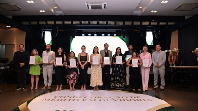 acre-realiza-solenidade-de-m-rito-em-ouvidoria-par Pessoas em solenidade recebendo certificados em um palco, com bandeiras e logotipos institucionais ao fundo.