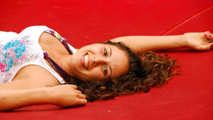 Grupo de 16 atletas de karatê posando em tatame, vestindo quimonos brancos e faixas coloridas, em ginásio esportivo.