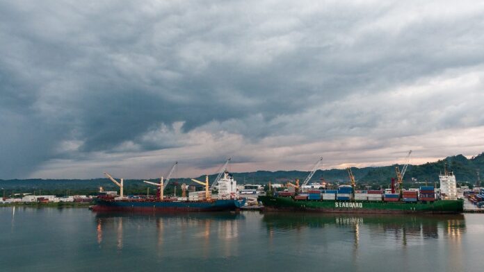 Vista aérea de navios de carga atracados em terminal portuário movimentado com grandes guindastes e contêineres.