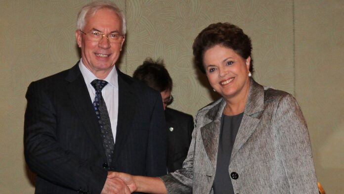 Fachada de um prédio bombardeado com escombros espalhados e fumaça ao fundo, em cenário de destruição urbana na Ucrânia.