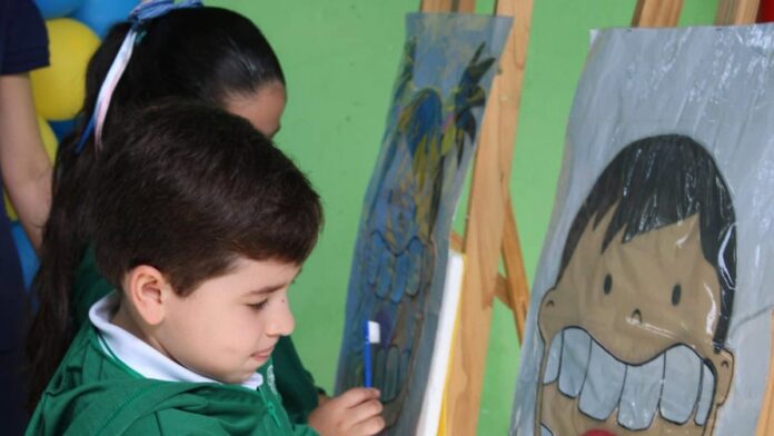 Dentista examina os dentes de uma criança em uma escola, com outros alunos aguardando atendimento ao fundo.