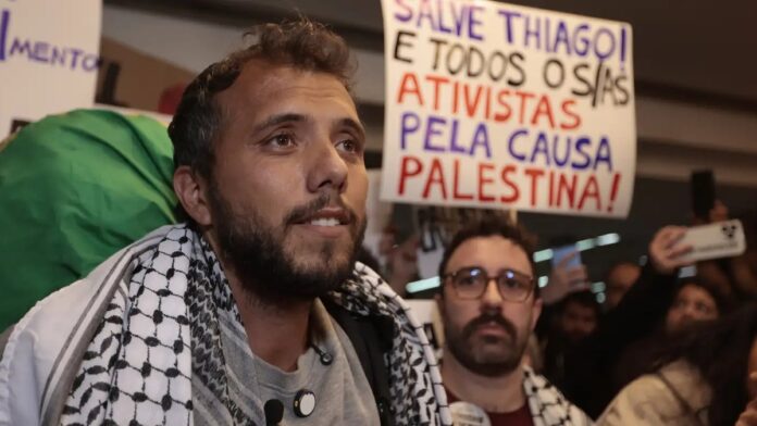 ativista-thiago-vila-deportado-da-argentina-ap-s-d Homem de barba e óculos em primeiro plano, com expressão séria, em meio a um ambiente urbano e movimento de pessoas.