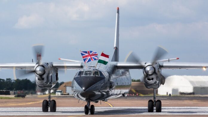 Destroços fumegantes de um avião militar em um campo aberto, com equipes de resgate trabalhando ao fundo.