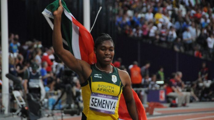Atleta de atletismo em pista de corrida, focada e em movimento, com fundo desfocado de estádio olímpico.