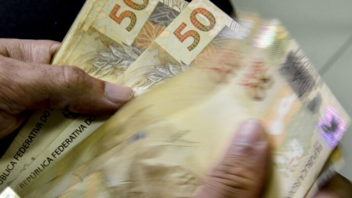 bolsa-fam-lia-caixa-paga-parcela-de-mar-o-a-benefi-1 Pessoa segurando um cartão do Bolsa Família próximo a uma agência da Caixa Econômica Federal.