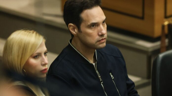 Jairinho sentado em tribunal, olhando para baixo, com advogados ao lado em uma sala de julgamento movimentada.