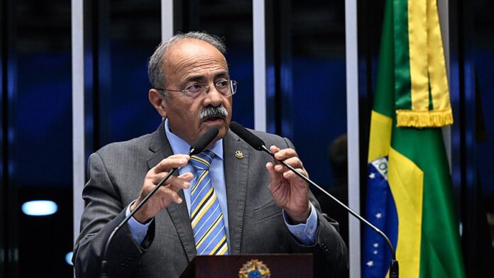 O senador Chico Rodrigues em entrevista, gesticulando e sorrindo, em um ambiente de escritório com bandeira ao fundo.