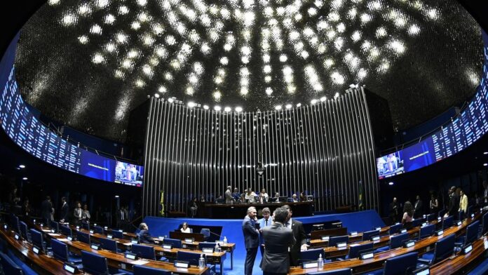 Fachada do Congresso Nacional, em Brasília, sob céu azul, com bandeiras do Brasil hasteadas ao vento.