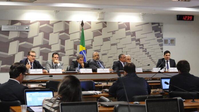 Parlamentares sentados em plenário durante sessão da CPMI, com foco em documentos sobre a mesa.