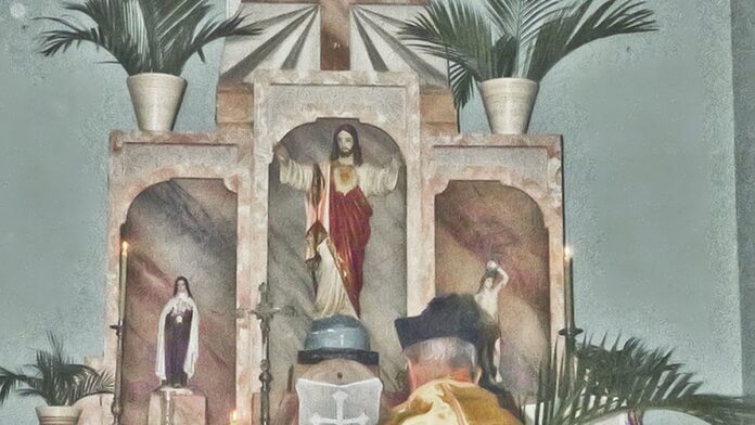 Mãos seguram ramos de palmeira entrelaçados em forma de cruz, com fundo desfocado em ambiente interno.