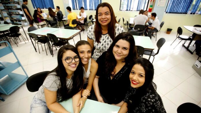 Estudante de enfermagem vestindo jaleco branco escreve em um caderno próximo a um notebook aberto sobre uma mesa.