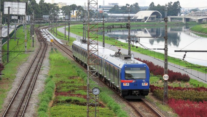 Entregador de uniforme laranja conduz uma bicicleta elétrica em rua movimentada da cidade com uma caixa térmica nas costas.