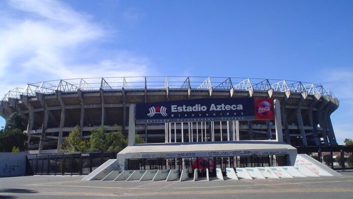 Vista aérea do Estádio Azteca iluminado, com gramado impecável e arquibancadas amplas sob céu ao entardecer.