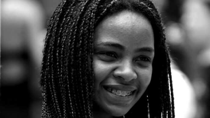 Estudante com mochila caminha em um campus universitário arborizado, segurando livros e olhando para um prédio clássico.