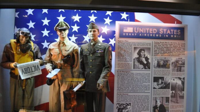 Helicóptero militar voando em baixa altitude sobre uma área arborizada com casas próximas.