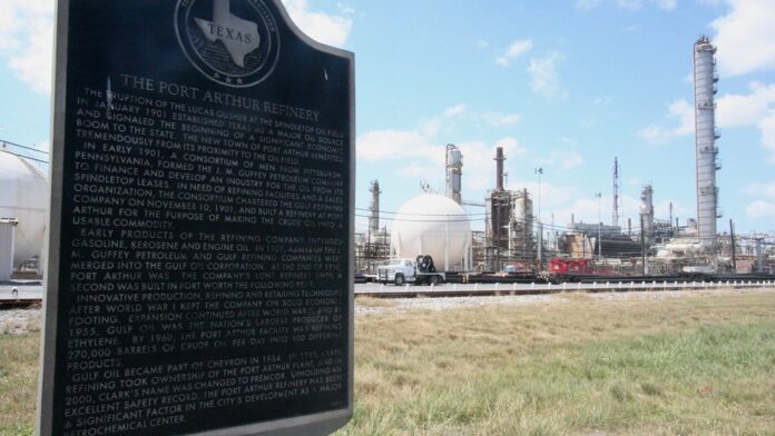 Coluna de fumaça escura e densa sobe de uma refinaria industrial, vista ao longe contra um céu aberto.