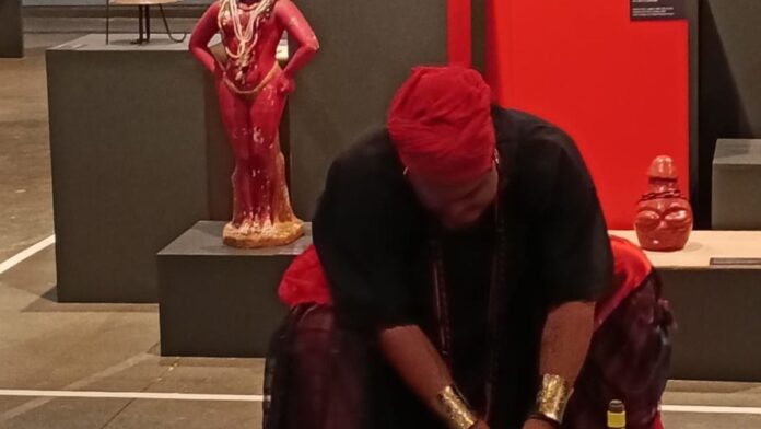 Escultura estilizada de Exu em madeira, com detalhes em búzios, exibida em pedestal no interior do Museu Afro Brasil.