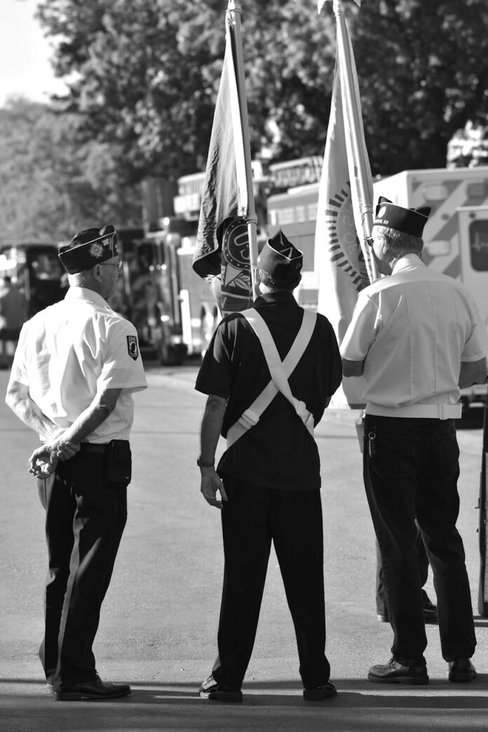 Três veteranos seguram bandeiras durante uma cerimônia patriótica ao ar livre em celebração ao serviço militar.