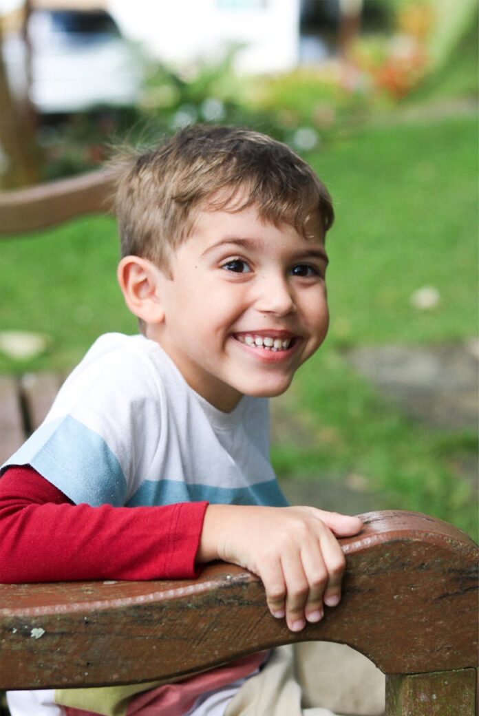 Menino brincalhão sentado em um banco de madeira ao ar livre, com um sorriso radiante.