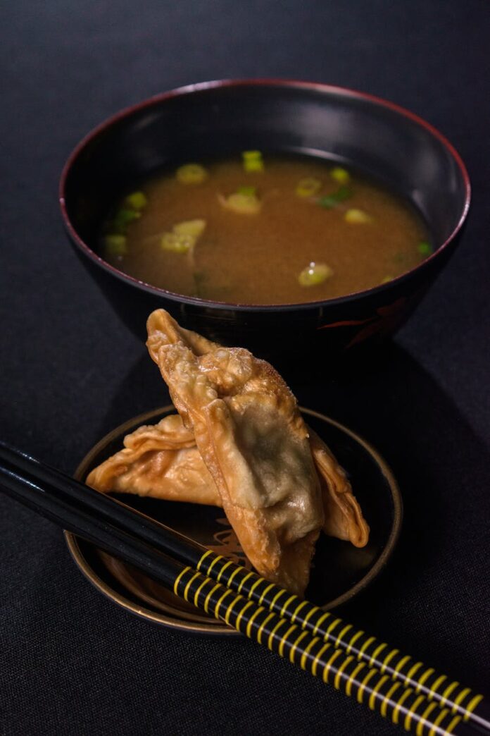 Uma visão de perto de sopa de missô com bolinhos de gyoza e hashis em uma superfície escura.