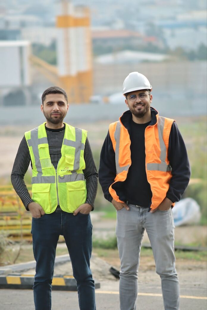 Dois operários da construção civil com equipamentos de segurança em um canteiro de obras ao ar livre, demonstrando trabalho em equipe.