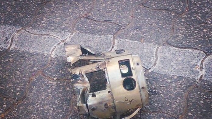 fragmento-de-m-ssil-encontrado-no-sul-de-israel-e- Fragmento metálico retorcido de um projétil caído sobre solo arenoso, com jovens observando a cena ao fundo.