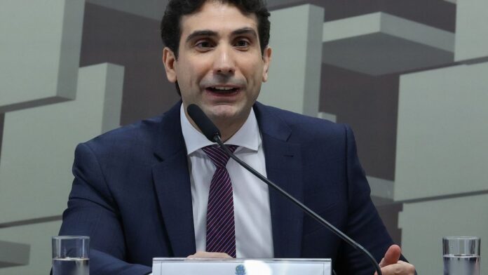 Secretário do Tesouro em entrevista coletiva, com gráficos de petróleo ao fundo.