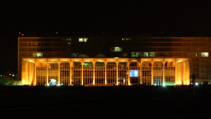 Vista aérea de uma cidade iraniana com fumaça ao fundo e silhueta de jatos militares cruzando o céu ao entardecer.