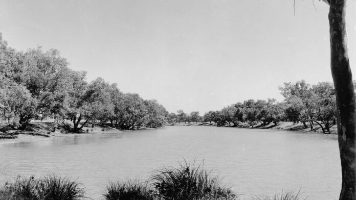 Três mulheres sorridentes sentadas à beira de um poço natural na Austrália, cercadas por rochas e vegetação rasteira.