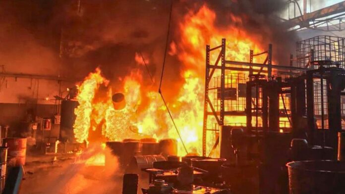 inc-ndio-de-grandes-propor-es-atinge-distribuidora Chamas altas e densa coluna de fumaça negra saindo de galpão industrial, com bombeiros combatendo o fogo no local.