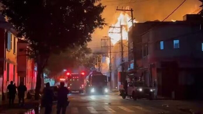 Chamas intensas e densa fumaça preta saem de um galpão industrial urbano durante incêndio.