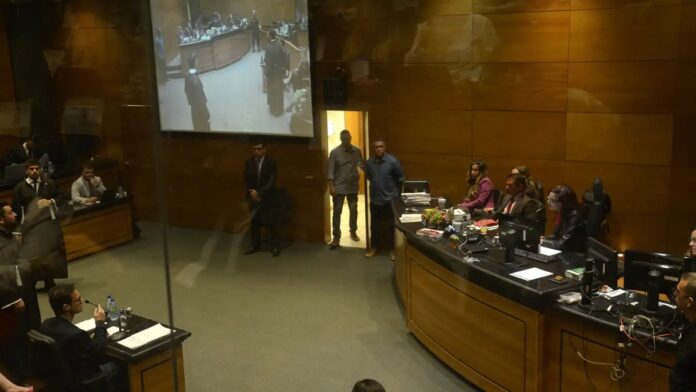 Monique Medeiros e Dr. Jairinho sentados lado a lado em tribunal, ambos com semblante sério e vestidos formalmente.