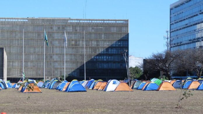 Policiais fardados em patrulhamento urbano, com viaturas e equipamentos táticos ao fundo.