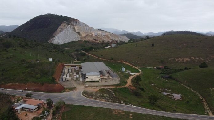 Vista aérea de uma grande mina a céu aberto com caminhões de transporte próximos a encostas rochosas expostas.