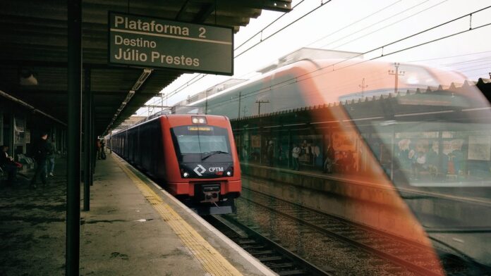 Trem moderno circulando em via férrea urbana com passageiros embarcando em plataforma de estação.