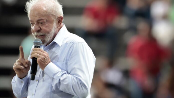 Lula em pronunciamento oficial, gesticulando com semblante sério, em frente a microfones e bandeira do Brasil.