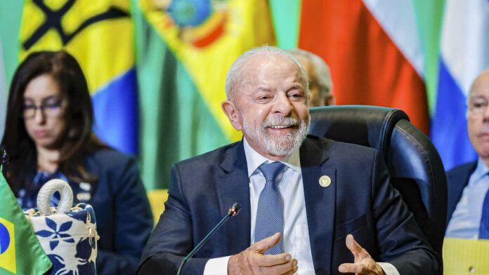 Lula discursa em cúpula da Celac, com bandeiras de países latino-americanos ao fundo e líderes em volta de uma mesa.