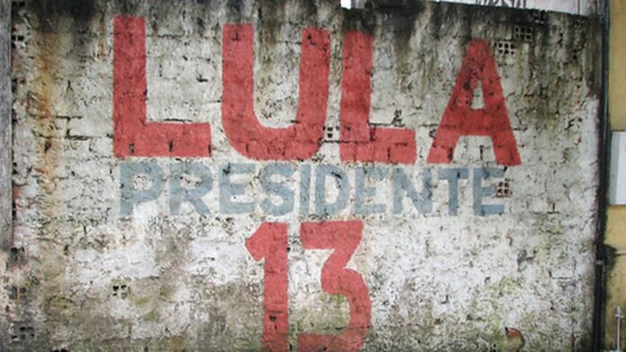 lula-lidera-inten-es-de-voto-contra-fl-vio-bolsona Luiz Inácio Lula da Silva discursa em um palco com microfones à frente e uma bandeira do Brasil ao fundo.