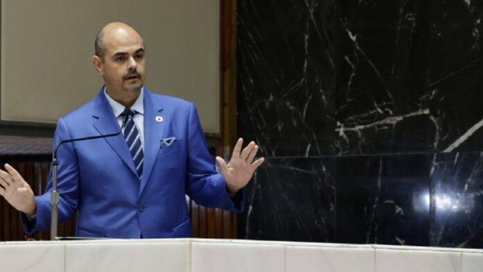 Mateus Simões discursa em cerimônia de posse na ALMG, diante de autoridades e bandeiras de Minas Gerais.
