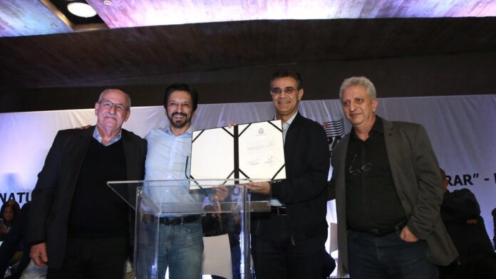 Mello Araújo em estúdio de TV, falando ao microfone com expressão séria, vestindo terno e camisa social branca.