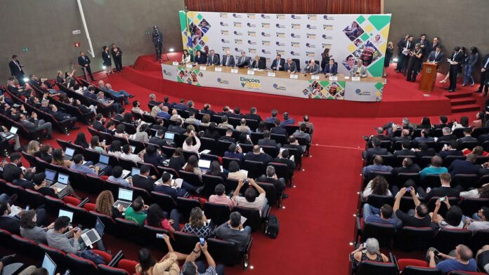 Dois homens em trajes formais apertam as mãos sobre uma mesa com documentos e bandeiras do Brasil ao fundo.