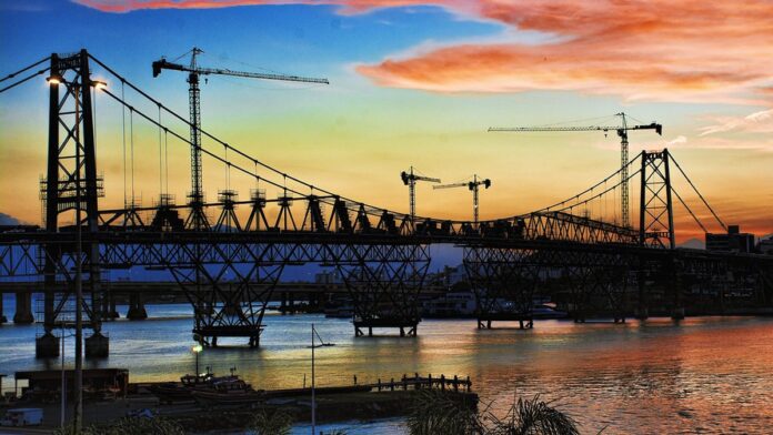 Estrutura em alvenaria da base policial em construção, com vigas expostas, andaimes e operários no canteiro de obras.