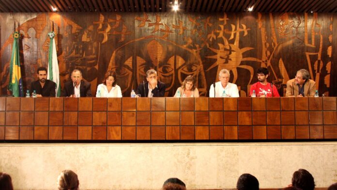 Servidores públicos em ambiente de escritório moderno, focados em telas de computador e interagindo entre si.