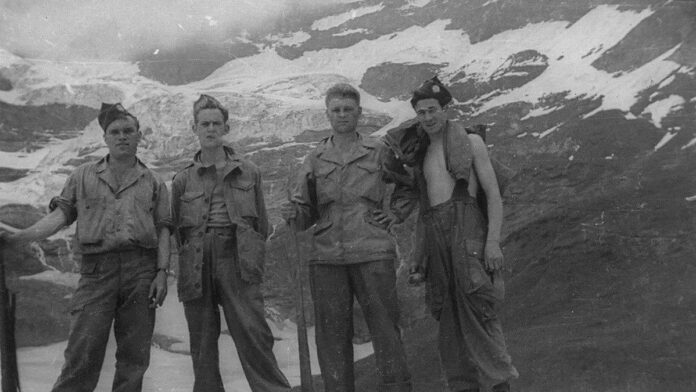 Pete Hegseth, de terno, discursa em um pódio com bandeiras dos EUA ao fundo em uma sala de imprensa oficial.