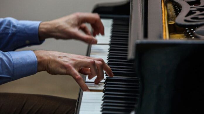 Músicos tocam instrumentos de sopro em um palco ao ar livre sob a luz do dia, com público ao fundo.