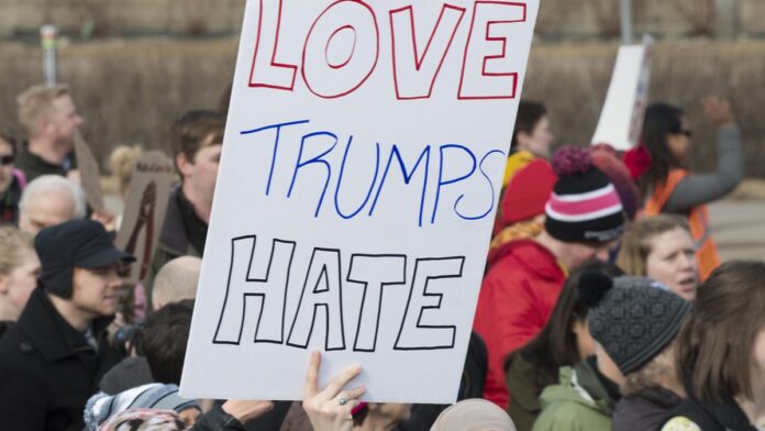 Multidão de manifestantes segura cartazes e bandeiras dos EUA em marcha urbana durante protesto contra Donald Trump.