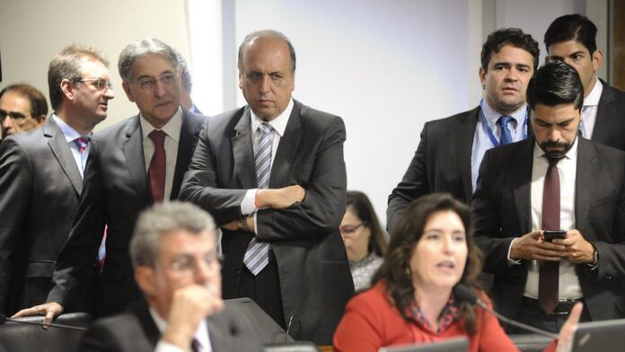 Funcionários do TRE-RJ organizam pilhas de cédulas eleitorais e urnas em uma mesa, em ambiente de trabalho administrativo.