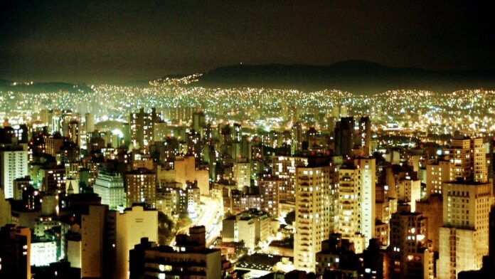 Mãos plantando uma muda em um vaso sobre escombros urbanos, com o horizonte de uma cidade cinzenta ao fundo.