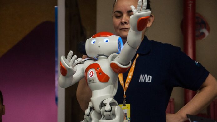 Robô humanoide prateado com sensores nos olhos gesticula em uma linha de montagem industrial iluminada.