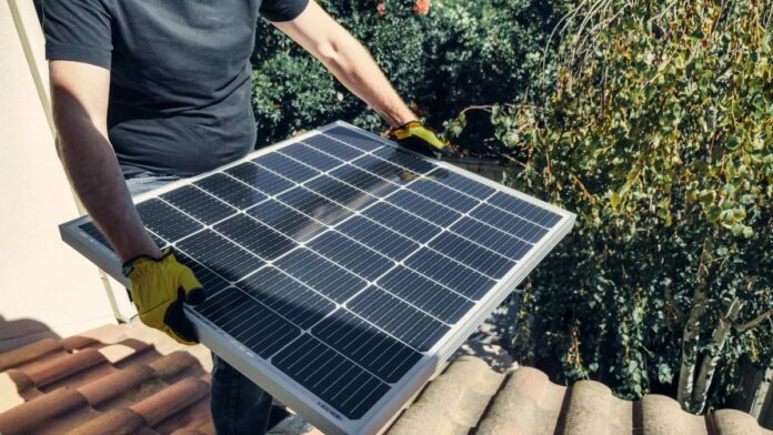 A worker installs a solar panel on a rooftop amidst lush greenery.